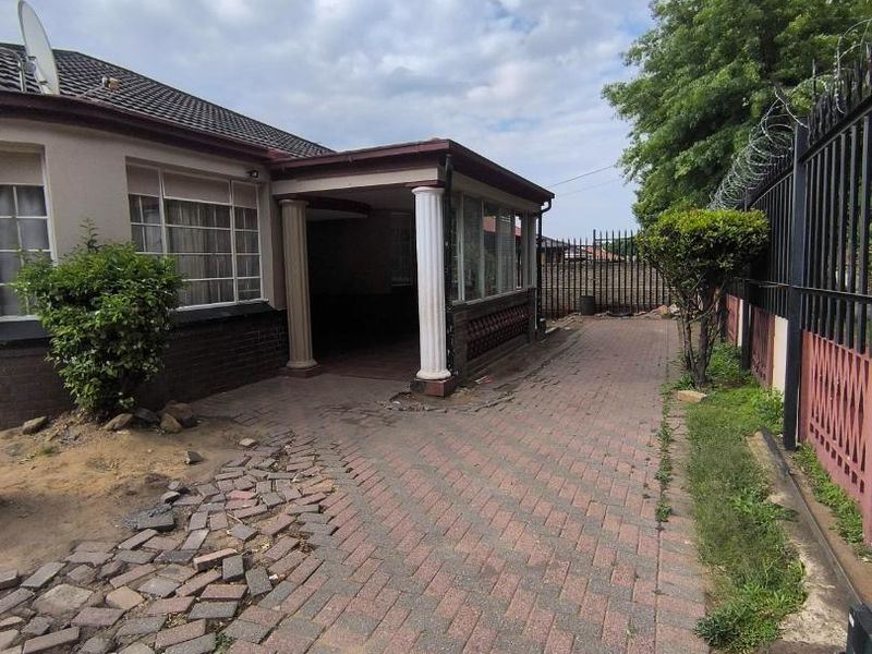 Front exterior view of the property featuring a paved driveway, covered entrance porch with decorative columns, and perimeter security fencing with razor wire.