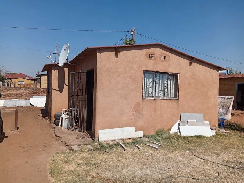 Rear view of a modest single-story dwelling featuring a simple plaster finish, metal security gate, and window bars.