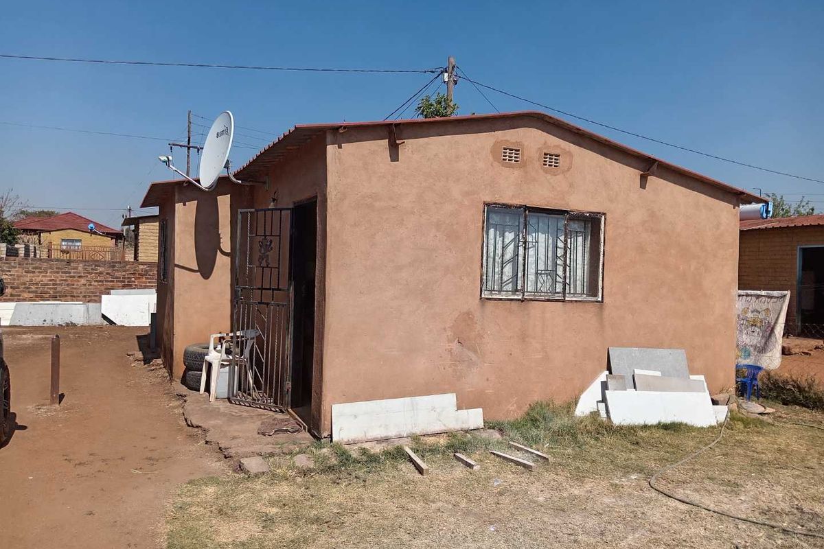 Rear view of a modest single-story dwelling featuring a simple plaster finish, metal security gate, and window bars.