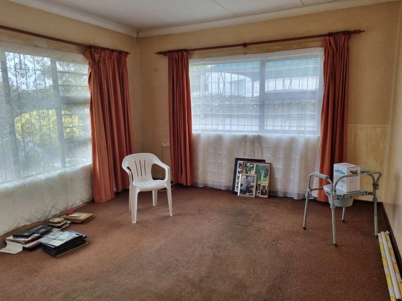 A spacious living area featuring large windows with security bars, carpeted flooring, and neutral walls in need of cosmetic updates.