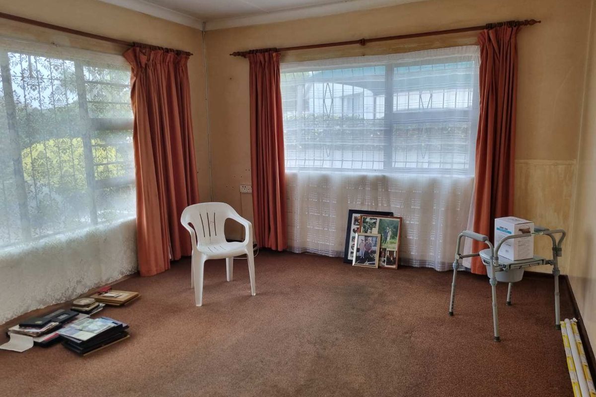 A spacious living area featuring large windows with security bars, carpeted flooring, and neutral walls in need of cosmetic updates.