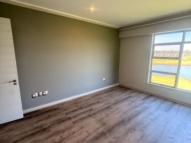 A bright, empty bedroom featuring modern laminate flooring, neutral wall tones, and a large window offering scenic water views.