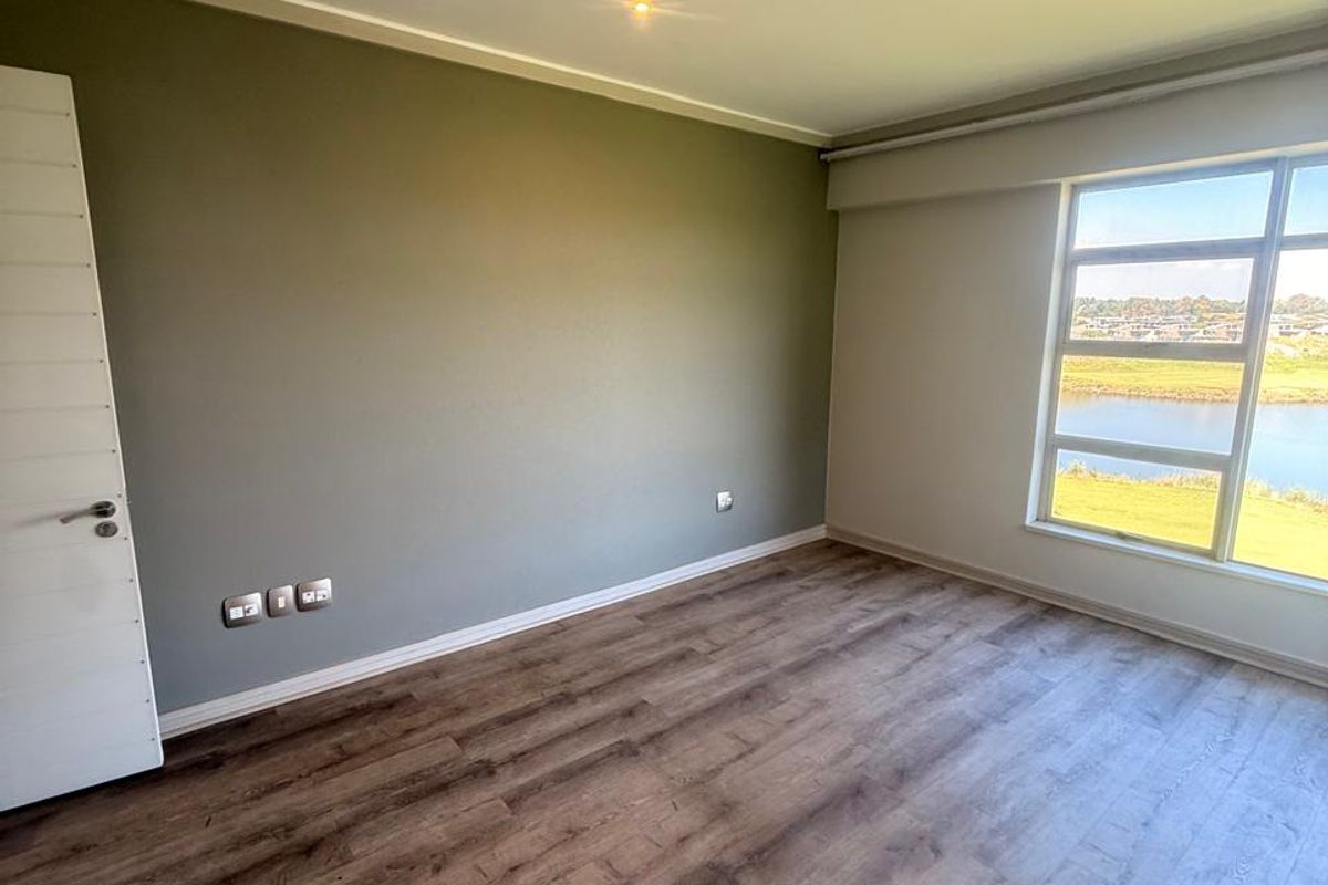 A bright, empty bedroom featuring modern laminate flooring, neutral wall tones, and a large window offering scenic water views.