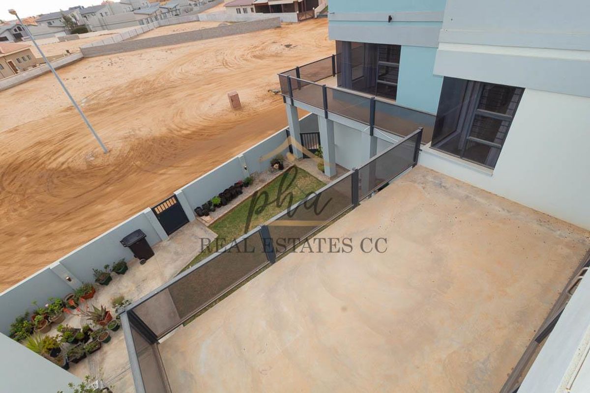 An elevated view of a modern residential property featuring a spacious paved patio area, a small garden patch, and glass-railed balconies.