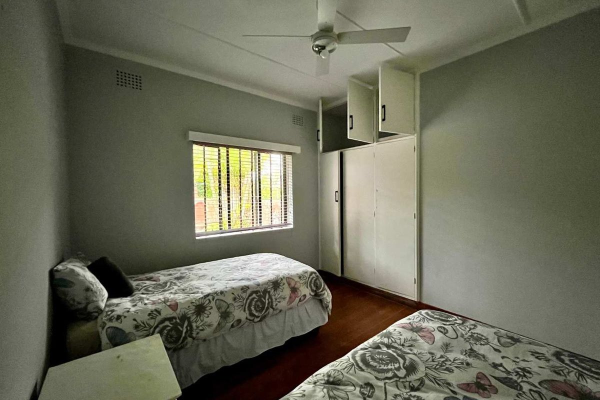 A bedroom featuring wooden flooring, built-in cupboards with overhead storage, a ceiling fan, and a window fitted with venetian blinds.