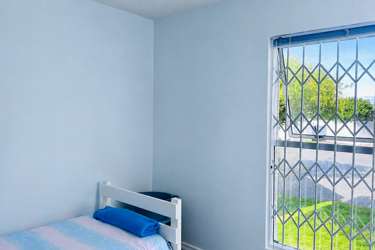 A bedroom featuring light blue walls and a window fitted with a metal security gate.