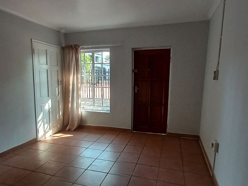 A bright, empty living room featuring tiled flooring, a timber entrance door, and a window fitted with security bars.
