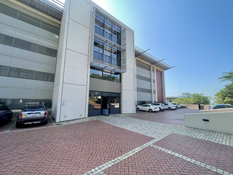Modern commercial office building exterior featuring a glass facade, aluminum sun louvres, and a paved entrance area.