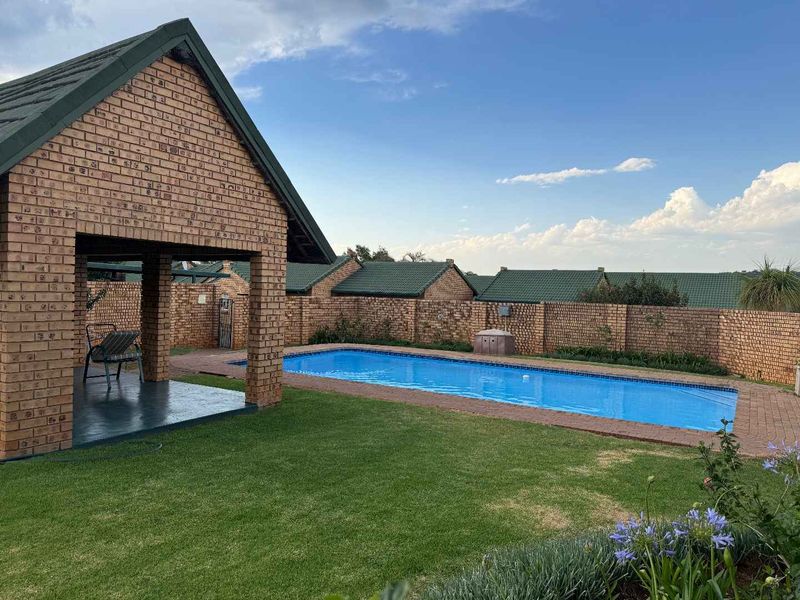 A well-maintained outdoor pool area featuring a rectangular swimming pool, brick paving, and a covered patio structure with a tiled roof.