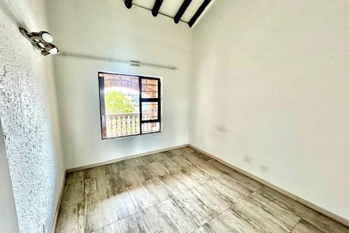 A bright, empty bedroom featuring high vaulted ceilings with exposed beams, wood-look tiled flooring, and a window overlooking the exterior balcony.