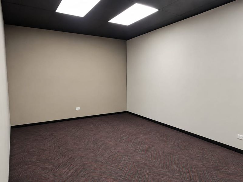 A clean, empty office space featuring dark carpet flooring, neutral walls, and recessed ceiling lighting, ready for professional use.