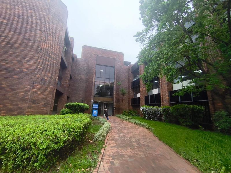 A multi-story office building featuring a classic brick facade, a paved pedestrian walkway, and well-maintained surrounding garden areas leading to the main entrance.