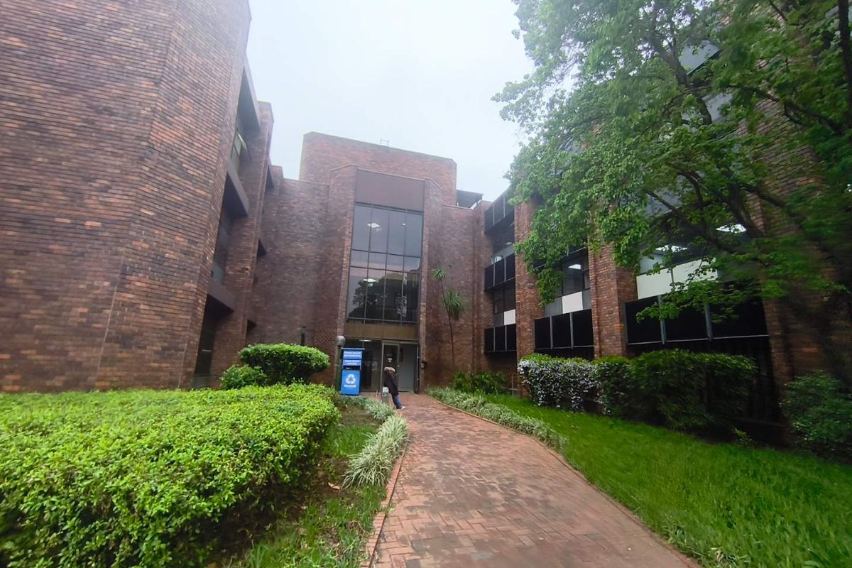 A multi-story office building featuring a classic brick facade, a paved pedestrian walkway, and well-maintained surrounding garden areas leading to the main entrance.
