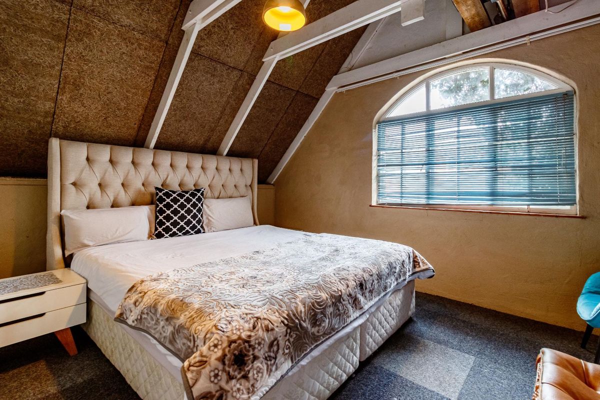A cozy attic-style bedroom featuring exposed timber rafters, a distinctive arched window, and textured wall paneling.