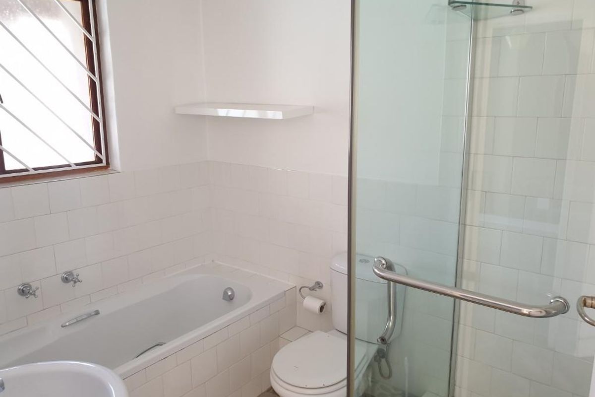 A functional bathroom featuring a built-in bathtub, a separate glass-enclosed shower, a toilet, and a window with security bars.