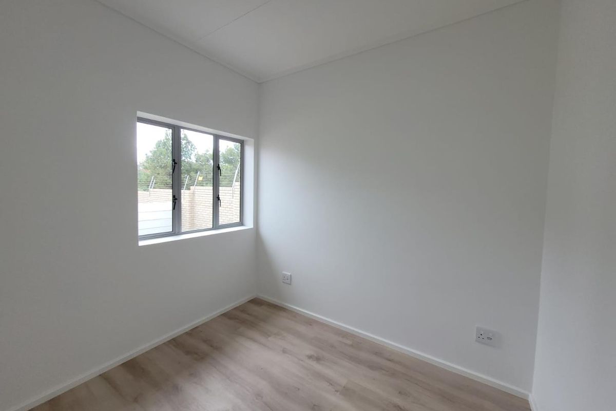 A bright, empty bedroom featuring neutral white walls, modern light wood-look flooring, and a window with aluminium frames.