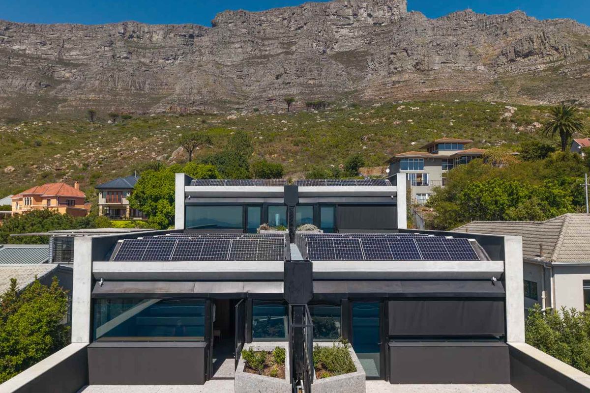 Aerial view of a modern residential property featuring solar panels, flat roof design, and integrated concrete planters against a mountain backdrop.