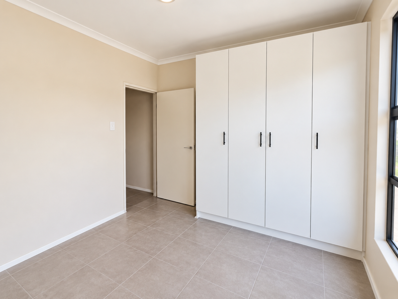 A bright, empty bedroom featuring neutral tiled flooring, a large window, and modern white built-in cupboards with black hardware.