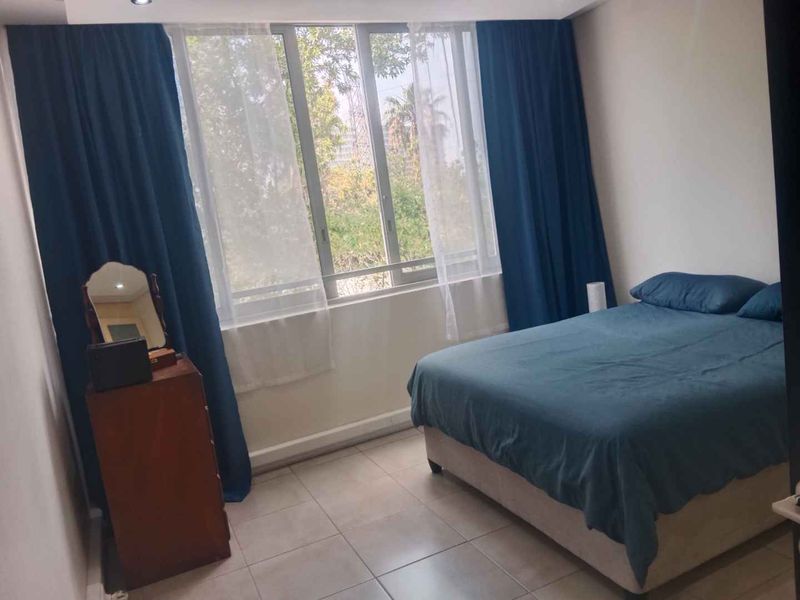 A bedroom featuring tiled flooring and a large window with curtains, providing natural light and a view of the surrounding greenery.