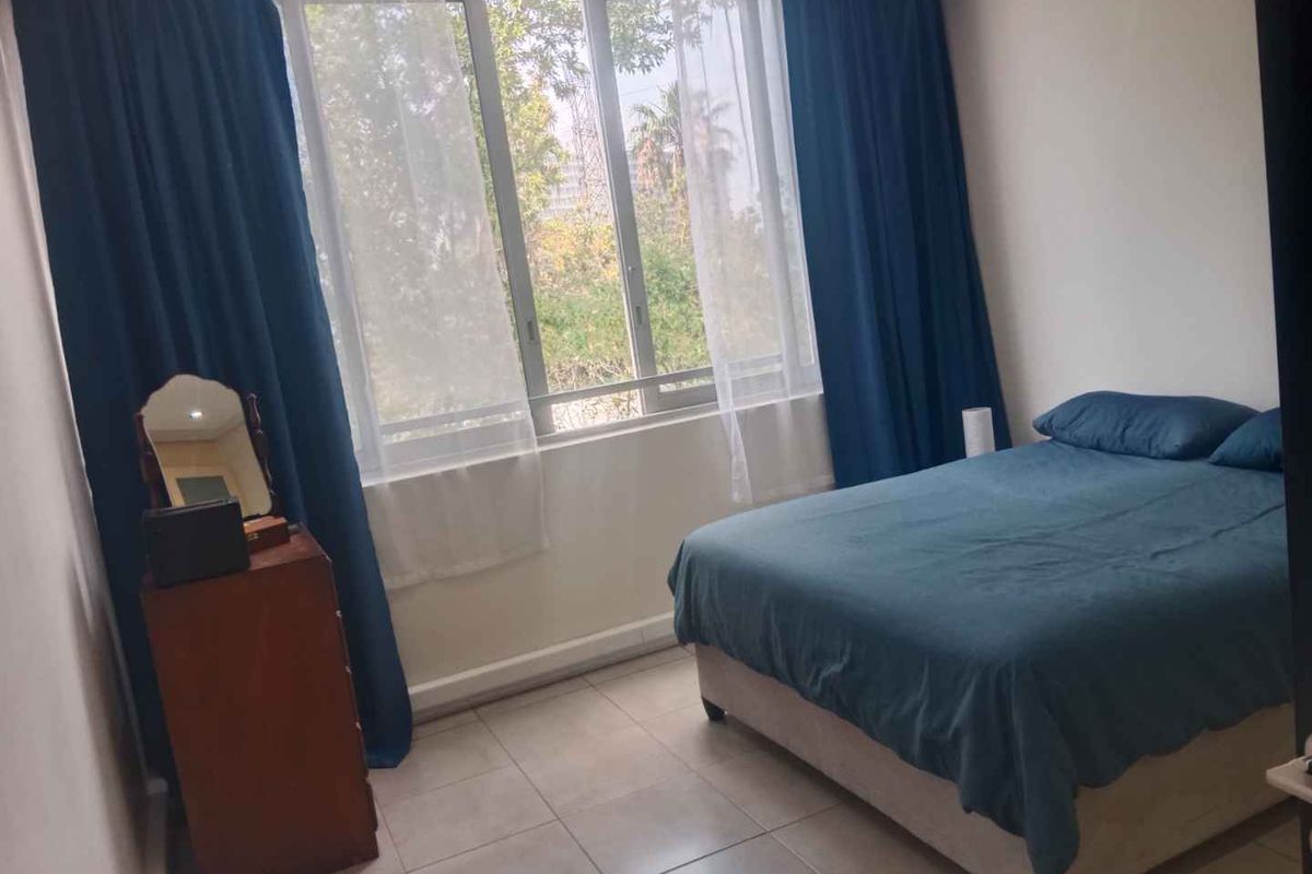 A bedroom featuring tiled flooring and a large window with curtains, providing natural light and a view of the surrounding greenery.