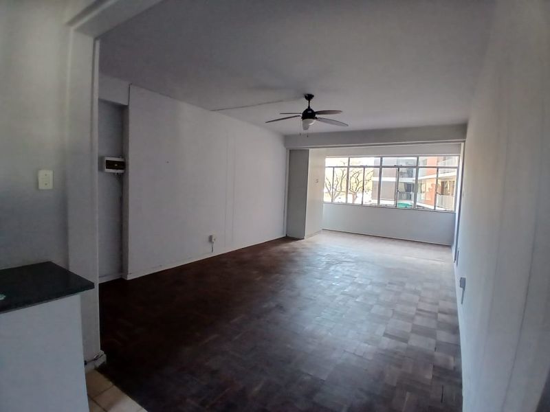 Spacious, empty living area featuring classic parquet flooring, a ceiling fan, and large windows providing natural light.