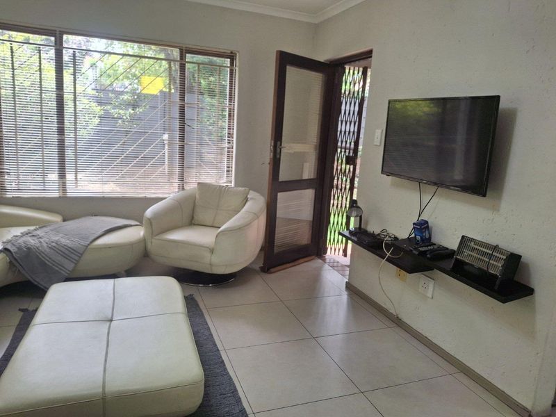 A living room featuring tiled flooring, a large window with venetian blinds, and a wall-mounted shelf with access to an exterior security gate.