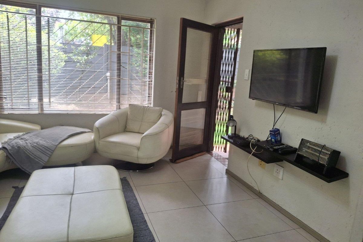 A living room featuring tiled flooring, a large window with venetian blinds, and a wall-mounted shelf with access to an exterior security gate.