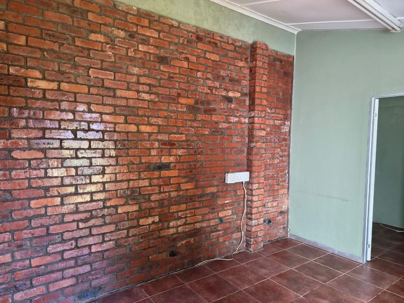 An empty retail space featuring an exposed brick feature wall, ceramic tile flooring, and a neutral painted wall with access to an adjacent room.