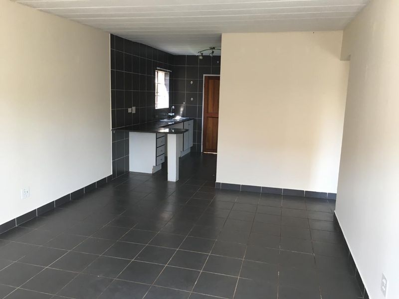 An open-plan living and kitchen area featuring dark floor tiles, neutral walls, and a functional kitchen with built-in cabinetry and a tiled splashback.