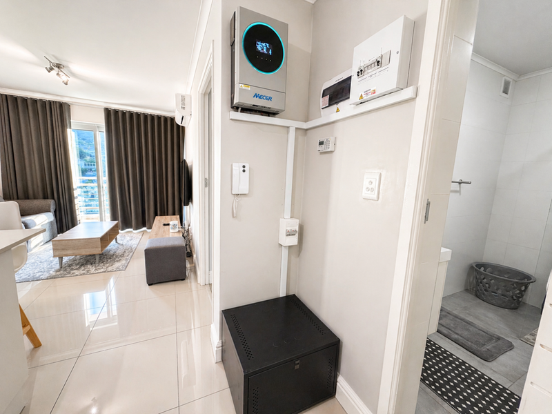 A clean hallway area featuring a wall-mounted inverter system, electrical distribution board, and intercom, with tiled flooring and access to adjacent living and bathroom spaces.
