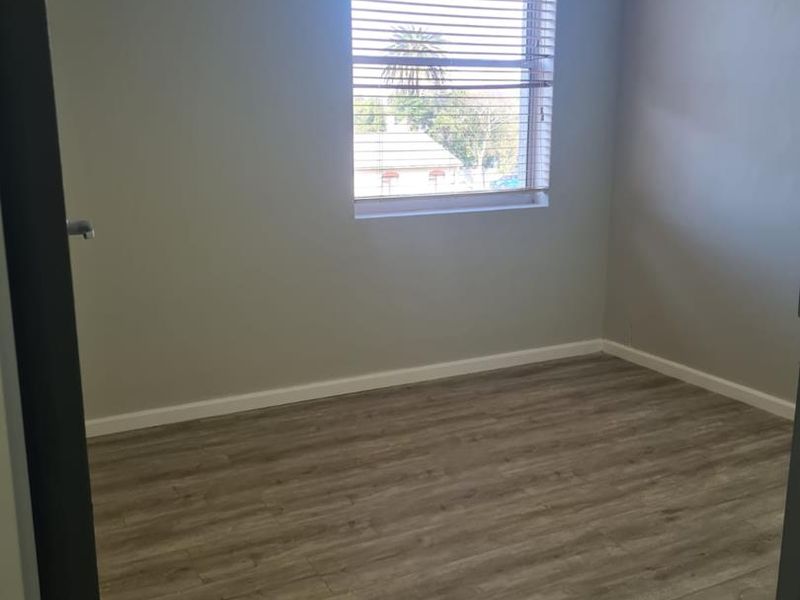 A clean, empty office space featuring modern laminate flooring, a drop ceiling, and a window with fitted venetian blinds.