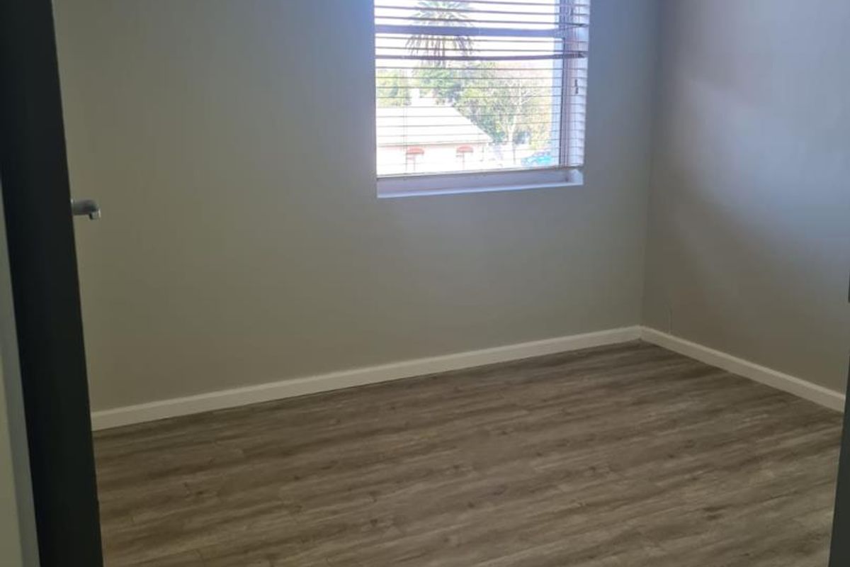 A clean, empty office space featuring modern laminate flooring, a drop ceiling, and a window with fitted venetian blinds.