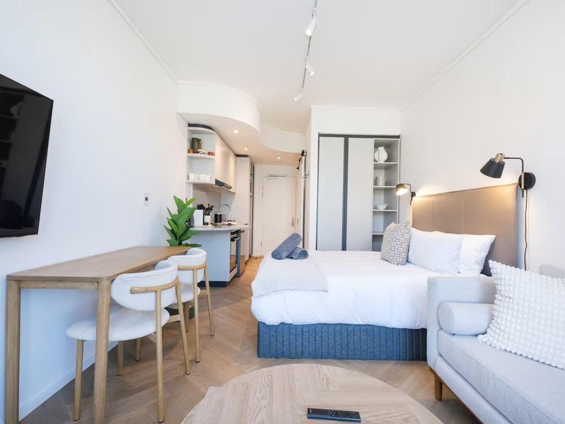 A modern open-plan studio apartment featuring a compact kitchenette, built-in wardrobe, track lighting, and stylish herringbone flooring.