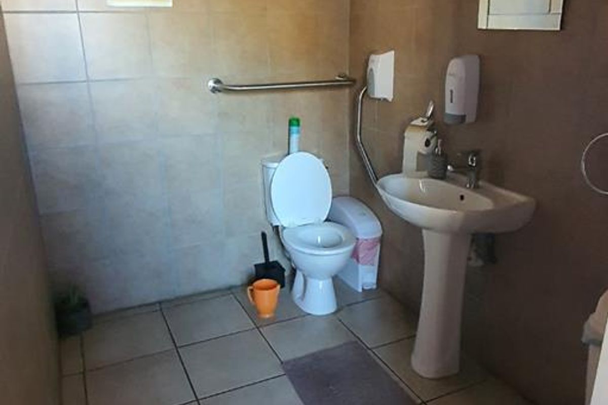 A functional commercial restroom featuring a pedestal washbasin, toilet, and wall-mounted grab rail with neutral tiled finishes.