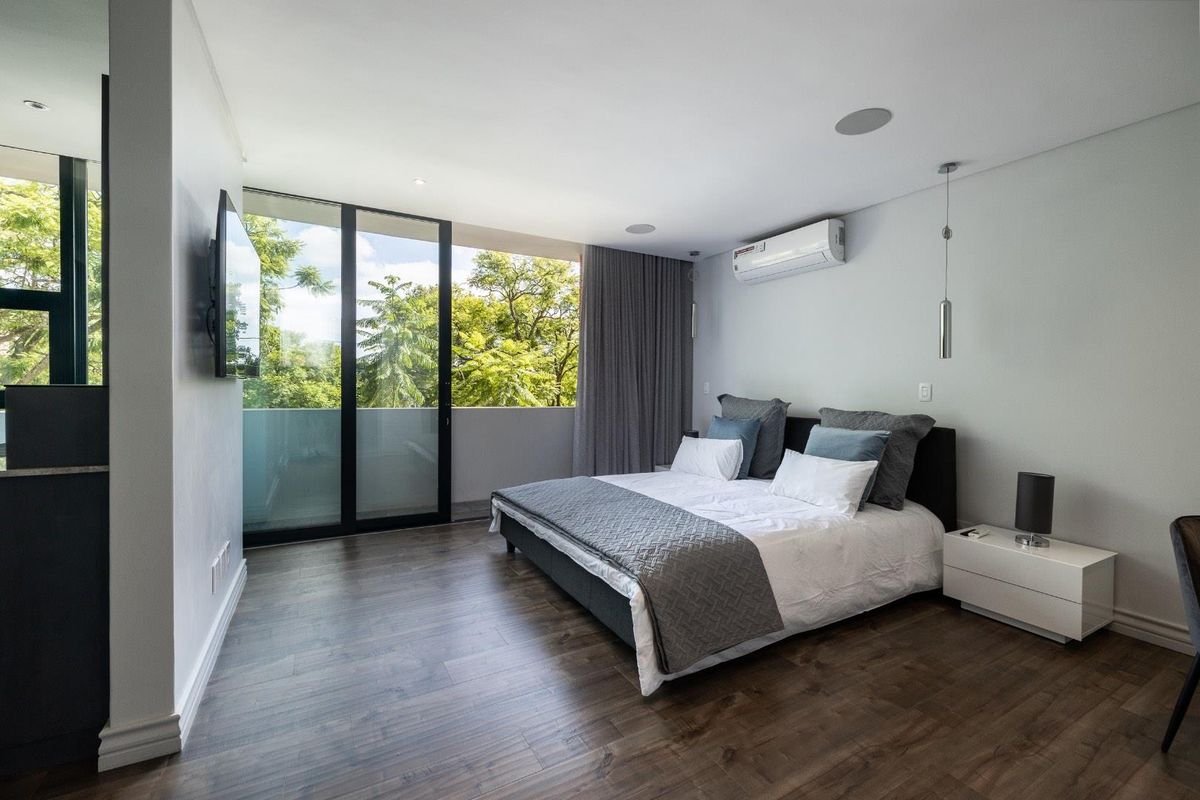 Modern bedroom featuring dark wood-look flooring, recessed ceiling lighting, integrated air conditioning unit, and large sliding glass doors opening to a private balcony.