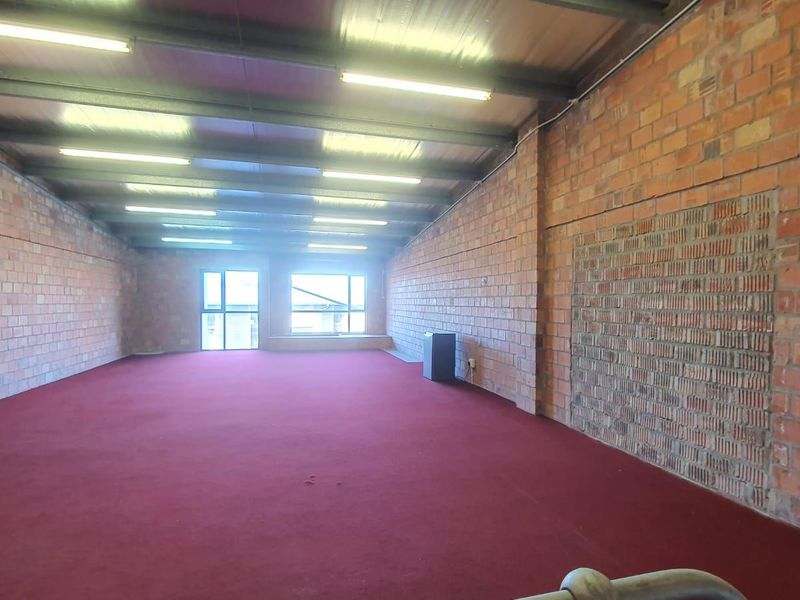 A spacious, open-plan industrial-style interior featuring exposed brick walls, high ceilings with exposed steel beams, and carpeted flooring.