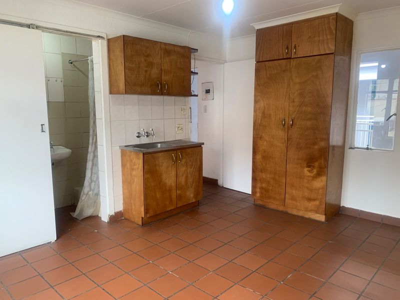 A compact kitchen area featuring built-in wooden cabinetry, a stainless steel sink, and tiled flooring, adjacent to a small bathroom space.