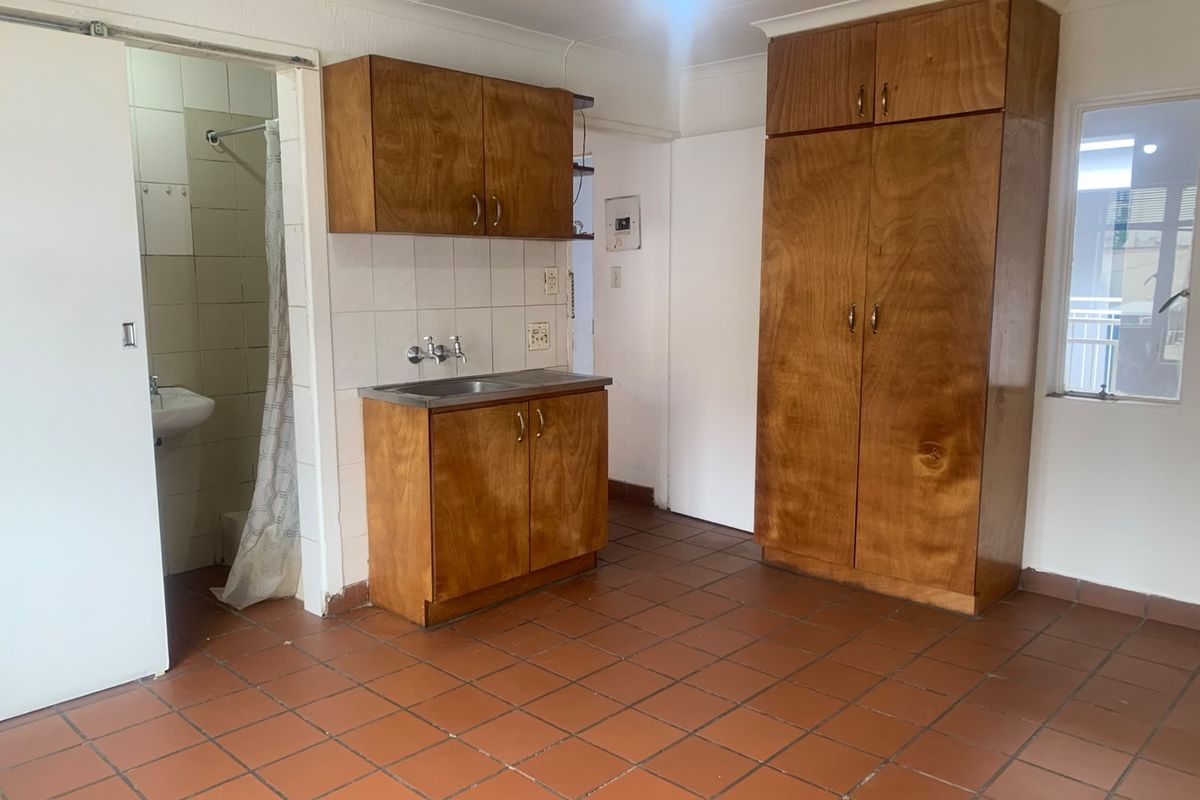 A compact kitchen area featuring built-in wooden cabinetry, a stainless steel sink, and tiled flooring, adjacent to a small bathroom space.