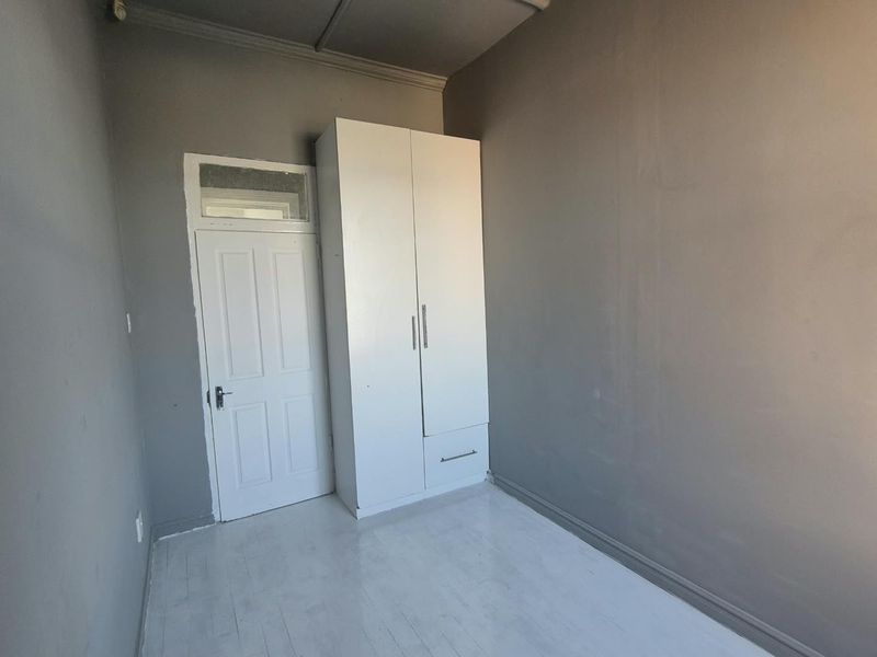 A simple, empty bedroom featuring grey walls, white painted wooden flooring, and a built-in white wardrobe.