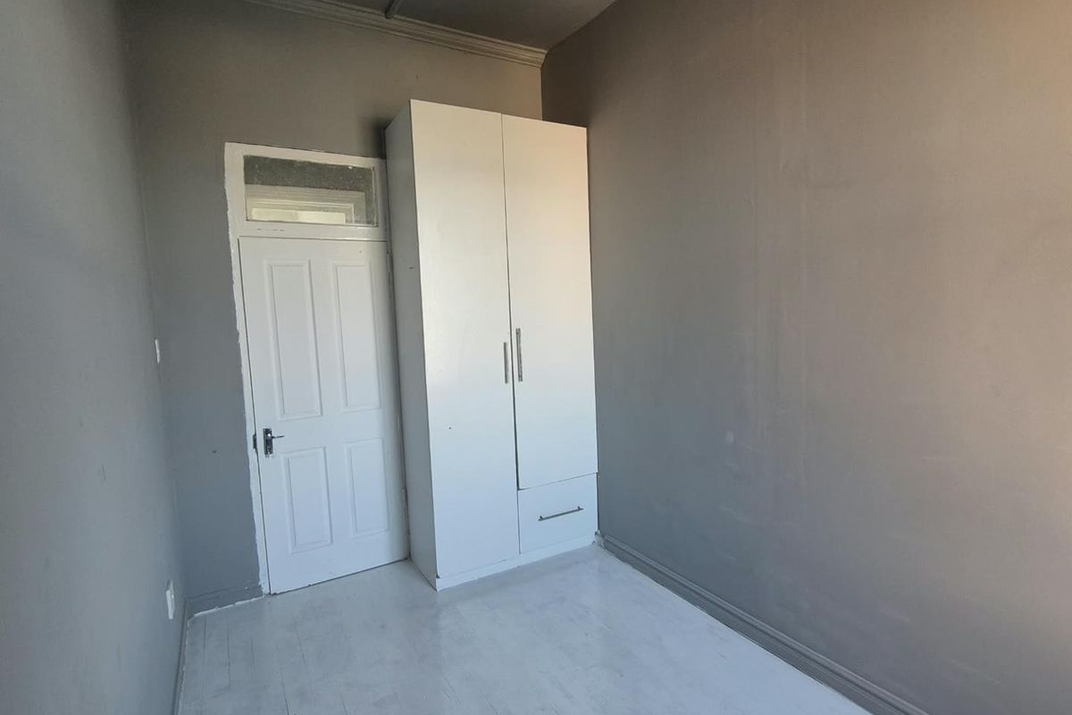 A simple, empty bedroom featuring grey walls, white painted wooden flooring, and a built-in white wardrobe.