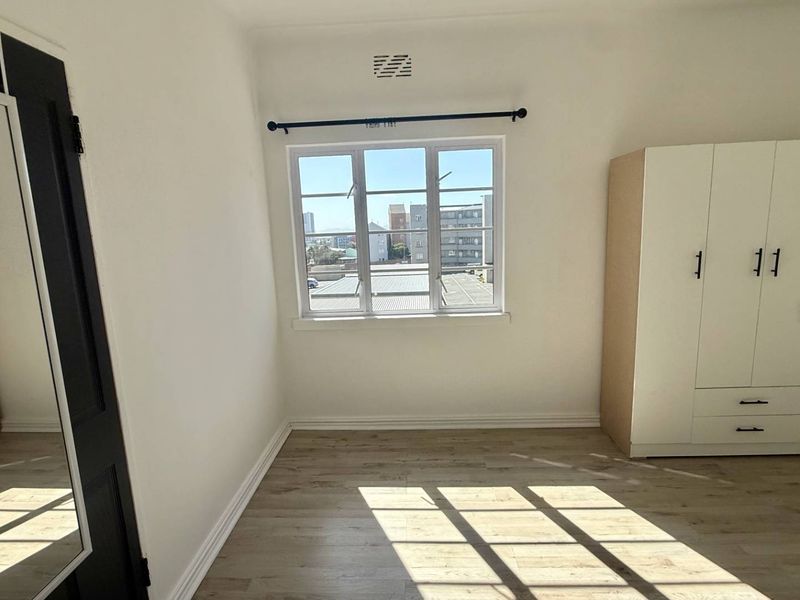 A bright, empty bedroom featuring light-toned laminate flooring, white walls, and a window with natural light.