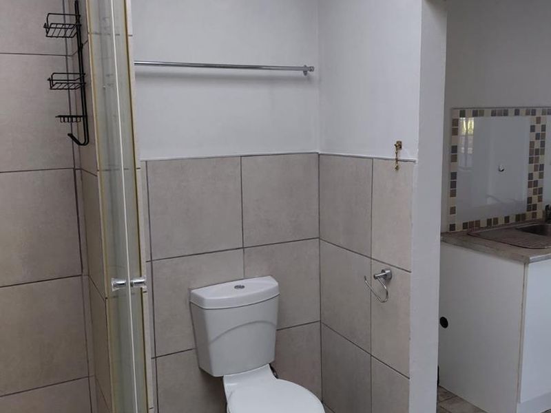 A functional bathroom featuring a toilet, shower enclosure, and neutral wall and floor tiling, with an adjacent view into a kitchen area.