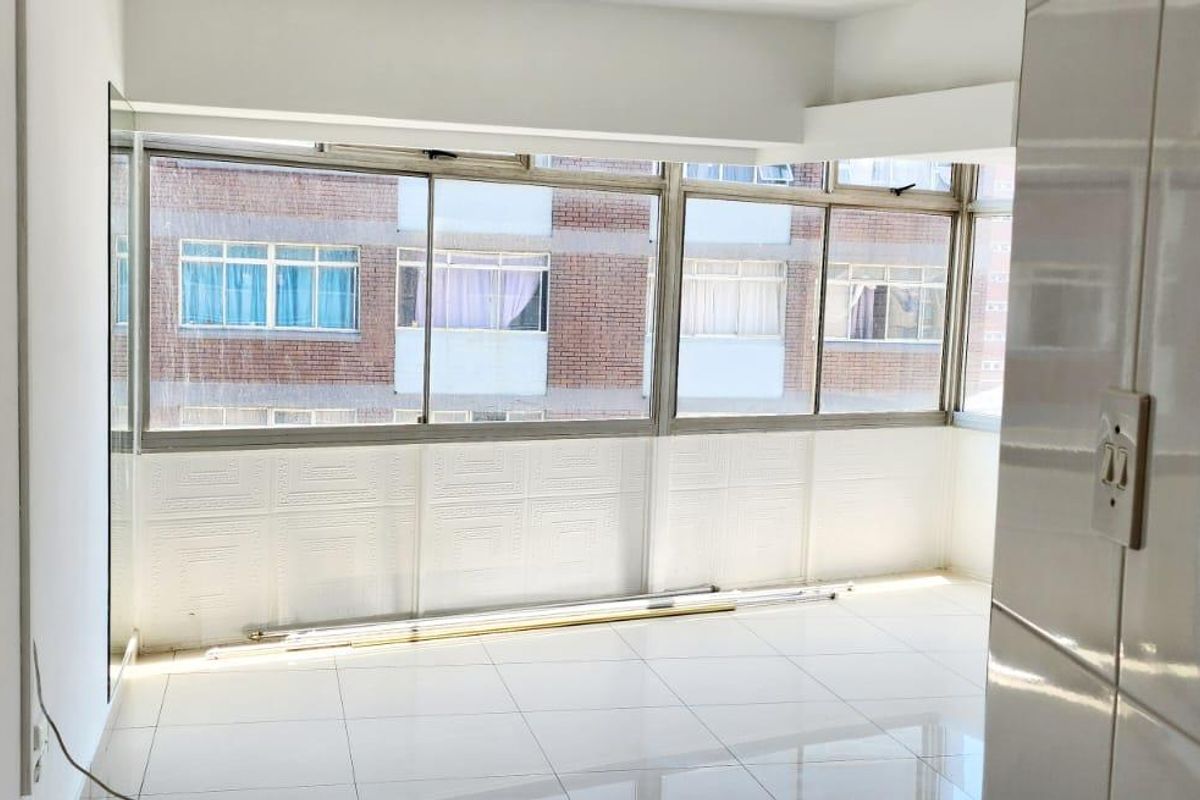 An empty, sunlit room featuring large sliding windows, white tiled flooring, and built-in wall paneling, offering a clean and versatile living space.