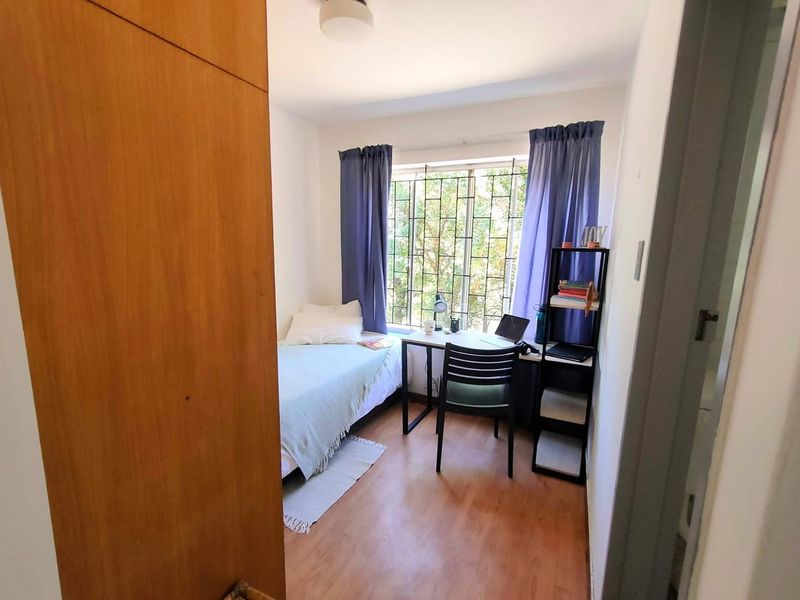 A compact bedroom featuring laminate flooring, a built-in cupboard, and a window fitted with security bars.