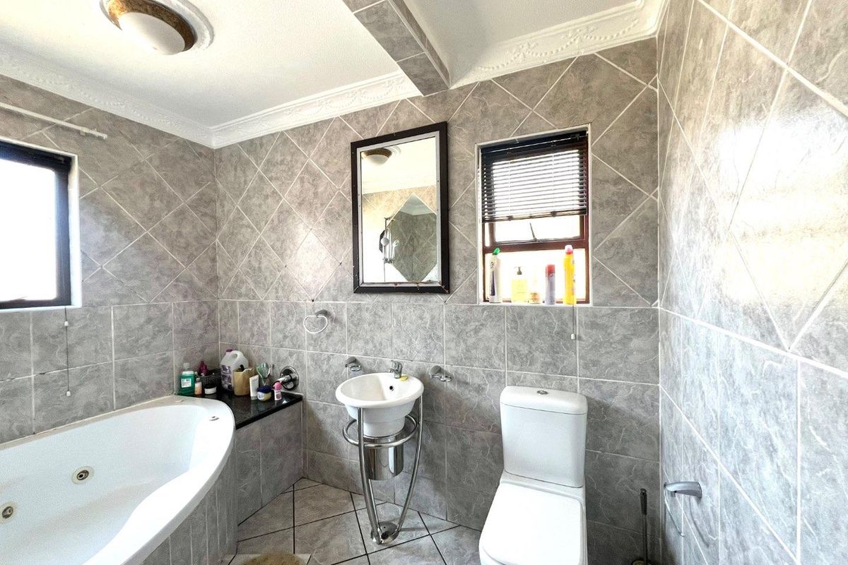 A compact bathroom featuring a corner jetted bathtub, pedestal basin, and toilet, finished with grey patterned wall and floor tiles.