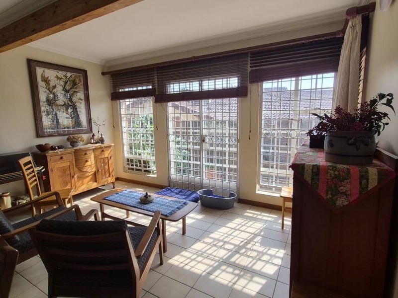 A sunlit living room featuring exposed timber ceiling beams, tiled flooring, and large windows with security bars, providing access to the exterior.