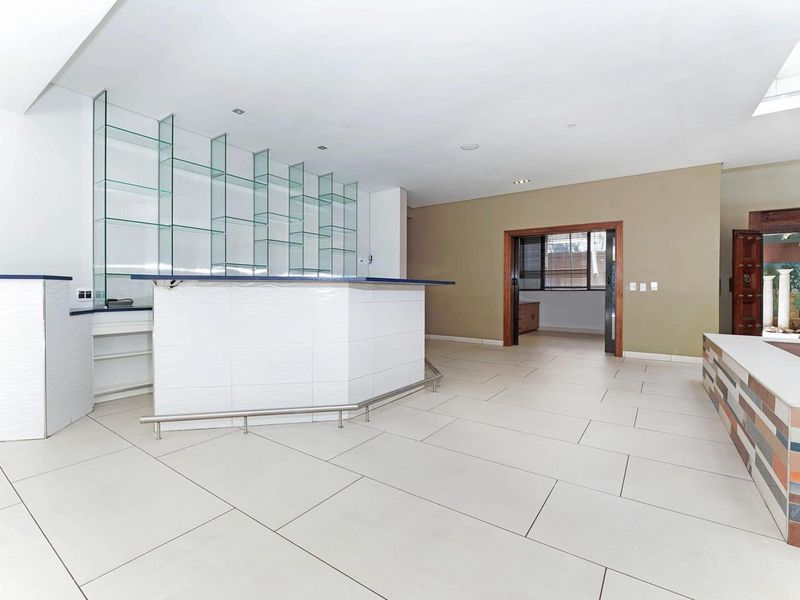 A spacious, empty bar area featuring a built-in counter, glass shelving, tiled flooring, and a skylight, offering a versatile entertainment space.
