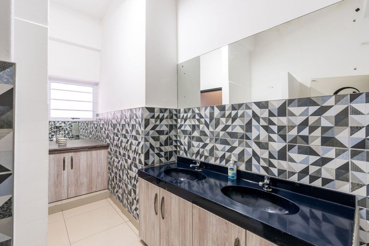 A clean, functional office bathroom featuring a double vanity with integrated sinks, patterned tiled backsplash, and a large wall-mounted mirror.
