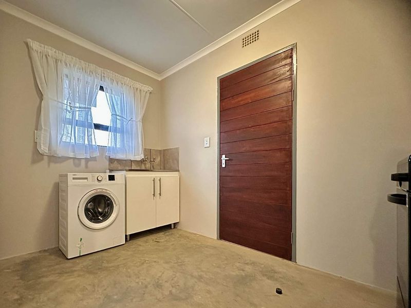 A functional laundry room featuring a built-in cabinet with sink, tiled splashback, and a solid timber door, with an unfinished concrete floor.