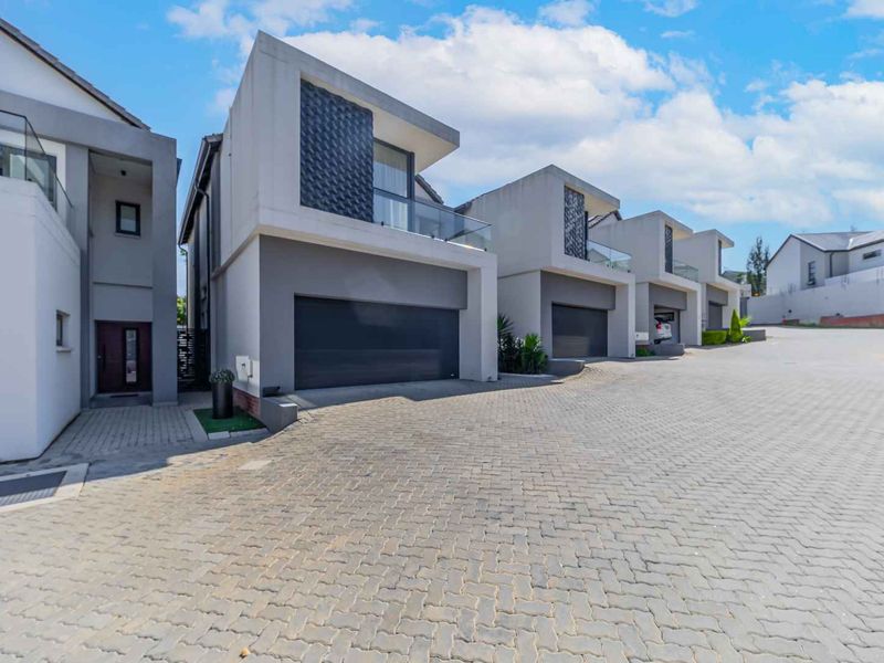 Modern cluster home exterior featuring a paved driveway, double garage, and upper-level balcony with glass balustrades in a contemporary residential estate.